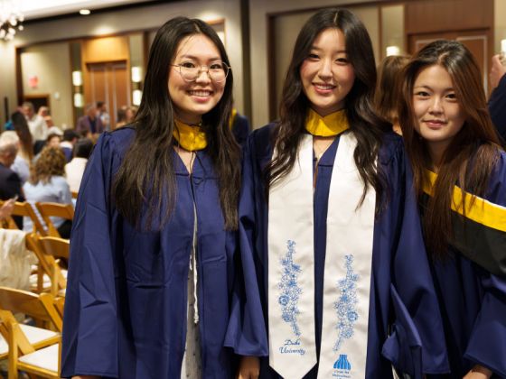 Three female graduates in blue gowns with yellow hoods pose together smiling at a reception; the center graduate wears a white Duke University stole from the Center for Multicultural Affairs.